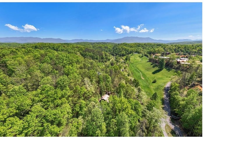 Aerial view reveals a pristine golf course nestled among rolling forested hills and distant mountain peaks.