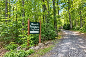 The property entrance is marked by a welcoming sign set within a serene, forested landscape with mature trees and a wellmaintained gravel driveway.
