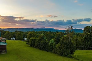 The property sits in a peaceful rural setting with lush green lawns, mature trees, and distant mountain ridges framing the horizon at sunset.