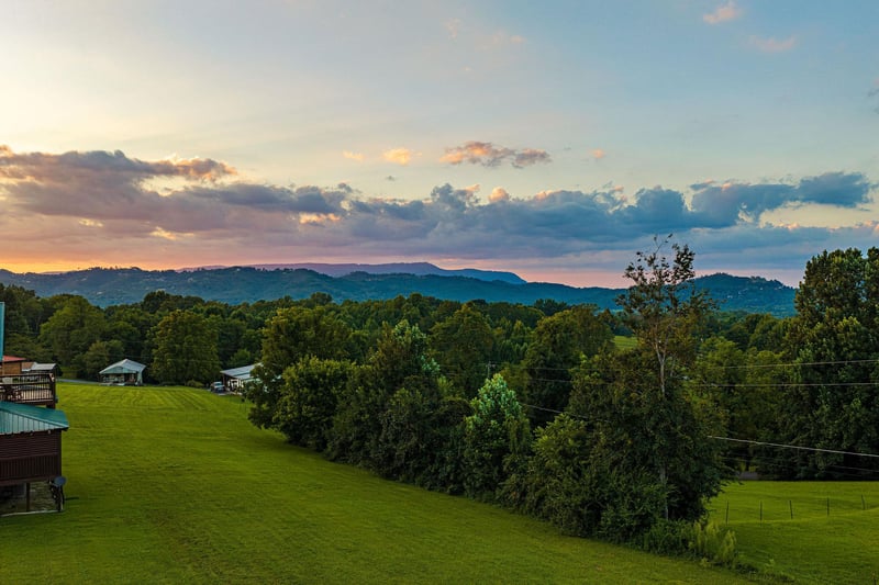 The property sits in a peaceful rural setting with lush green lawns, mature trees, and distant mountain ridges framing the horizon at sunset.