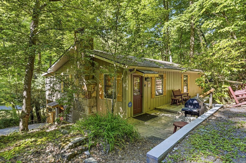 A charming woodland cabin nestled among towering trees, featuring a welcoming covered porch with seating and a BBQ grill on the deck.