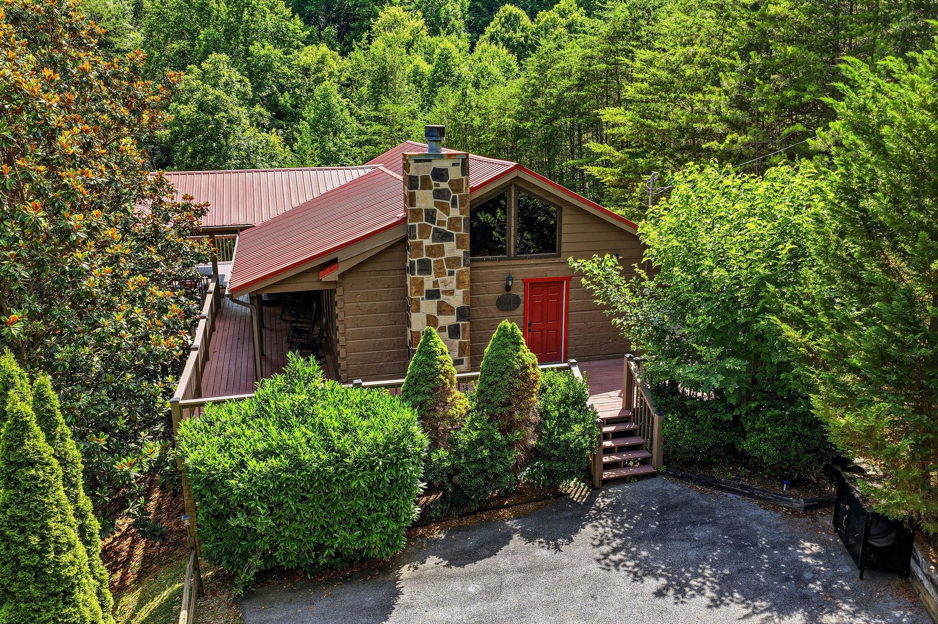 A charming mountain cabin with a distinctive stone chimney and red metal roof nestled in lush forest. The welcoming red door and wellmaintained landscaping create an inviting entrance to this seclude