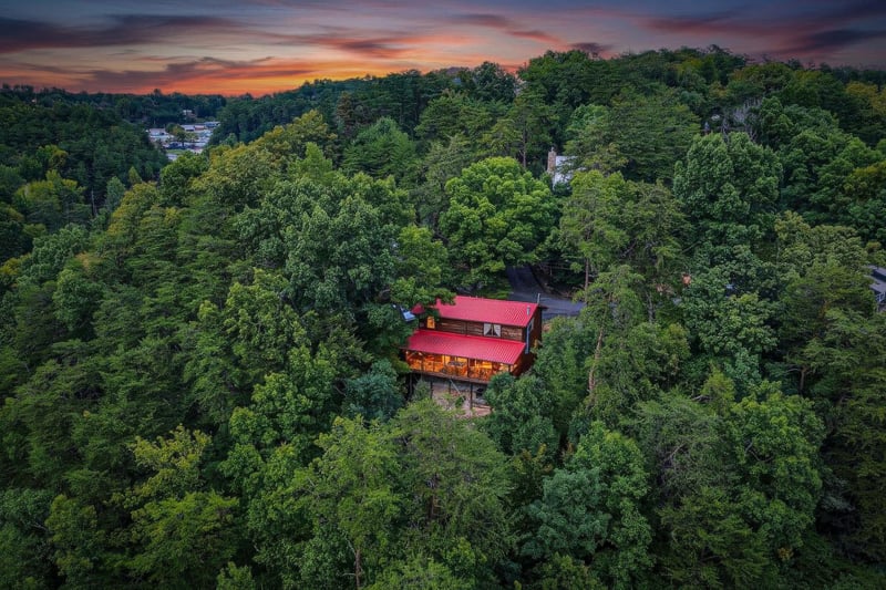 A striking cabin nestled among lush wooded hillsides, with a vibrant red roof standing out against the forest canopy as the sun sets over the peaceful mountain area.