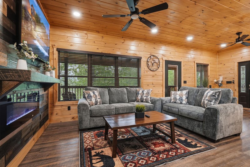 Sink into plush gray sofas by the stone fireplace in this cozy cabin living room, where rustic wood ceilings and warm lighting create your perfect mountain retreat.