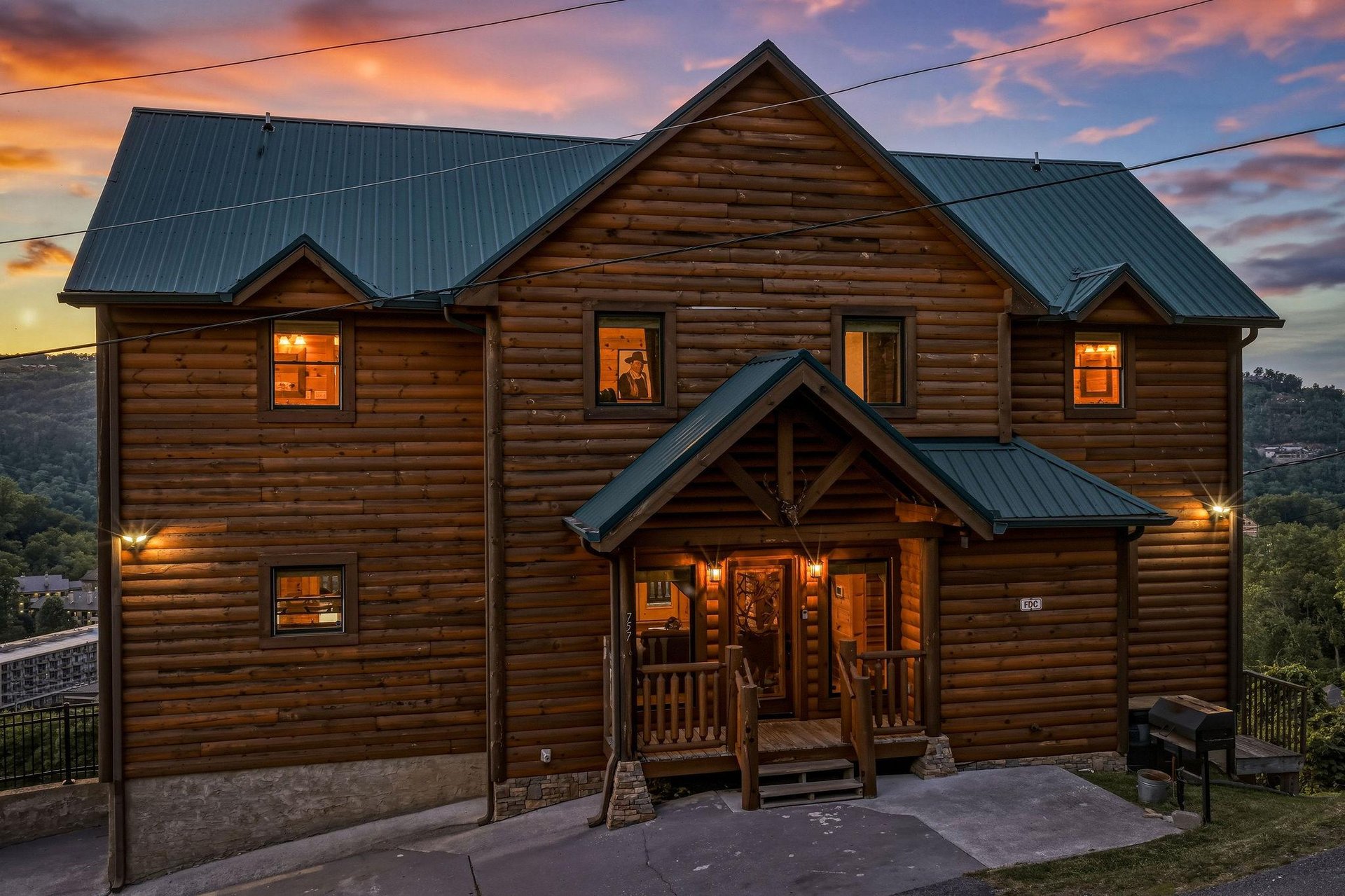 Stunning log cabin with warm lighting glows invitingly against a vibrant sunset sky, set among rolling hills in a peaceful mountain community.