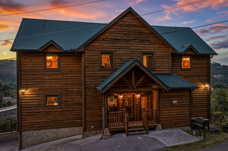 Stunning log cabin with warm lighting glows invitingly against a vibrant sunset sky, set among rolling hills in a peaceful mountain community.