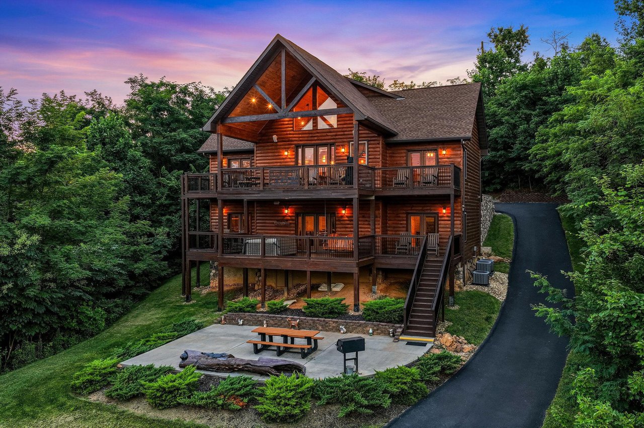 A charming log cabin retreat nestled among lush trees, featuring multilevel decks and a welcoming patio area perfect for evening gatherings as the sun sets.