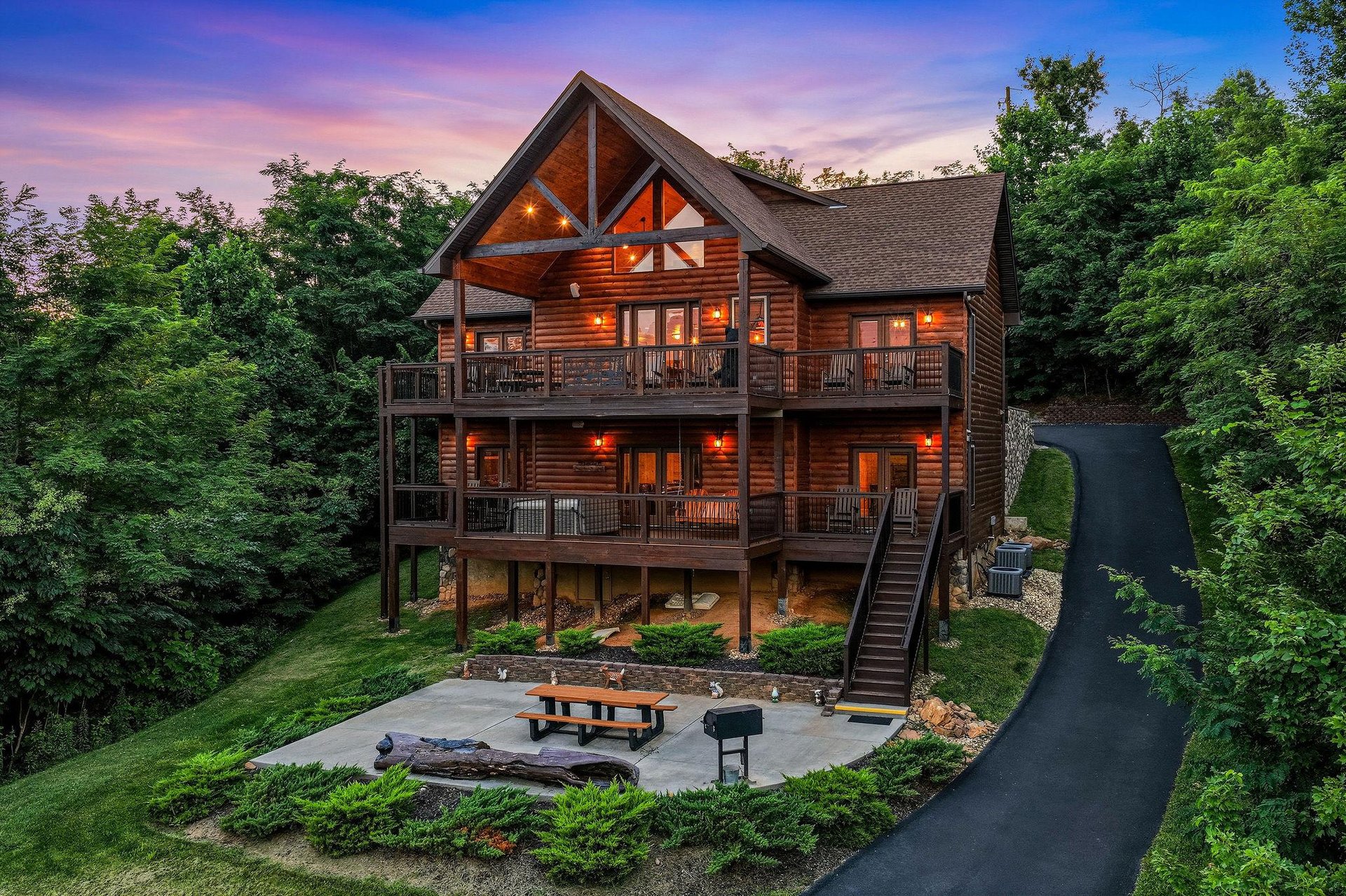 A charming log cabin retreat nestled among lush trees, featuring multilevel decks and a welcoming patio area perfect for evening gatherings as the sun sets.