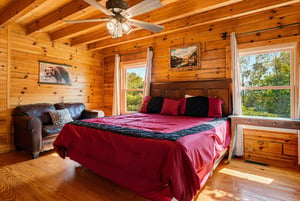Unwind in your cozy cabin bedroom with rustic knotty pine, a plush bed with vibrant linens, and sunlight streaming through large windows framing peaceful tree views.