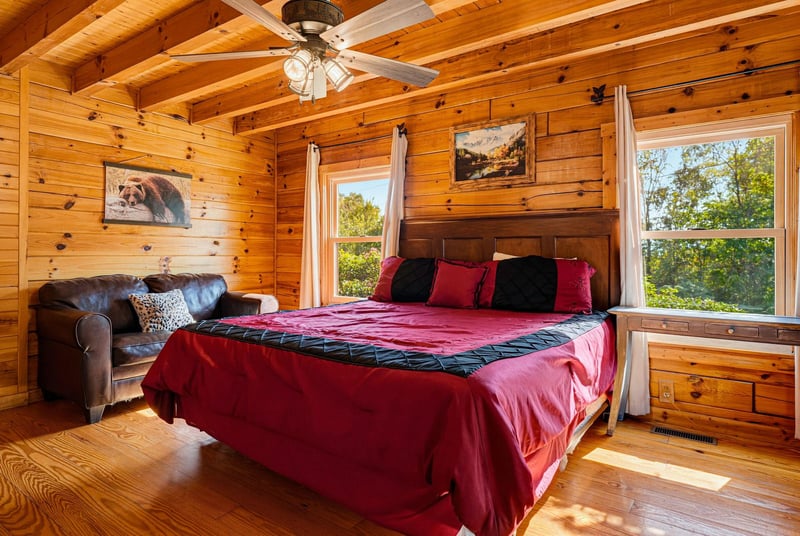 Unwind in your cozy cabin bedroom with rustic knotty pine, a plush bed with vibrant linens, and sunlight streaming through large windows framing peaceful tree views.