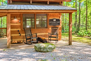 The charming facade of Little Bird Cabin welcomes guests with twin rocking chairs on a covered porch, surrounded by lush forest greenery and serene mountain solitude.