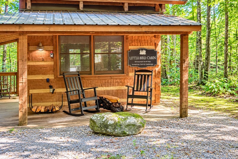 The charming facade of Little Bird Cabin welcomes guests with twin rocking chairs on a covered porch, surrounded by lush forest greenery and serene mountain solitude.