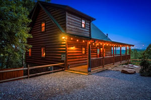 A welcoming mountain cabin awaits at twilight, with warm lighting and a spacious covered deck offering distant mountain views.