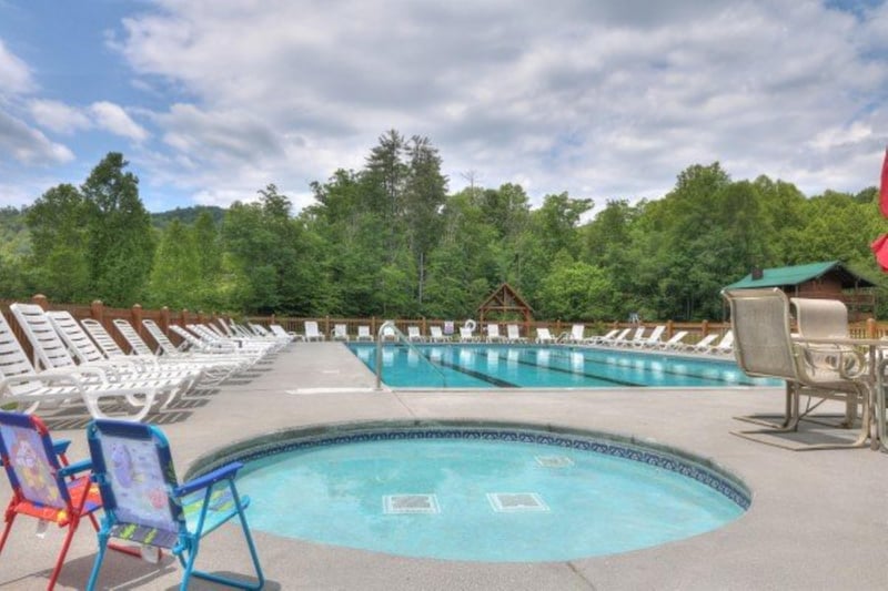 A spacious outdoor pool area surrounded by lush forests and mountains, featuring ample sun loungers and a relaxing hot tub, perfect for soaking up the peaceful mountain setting.