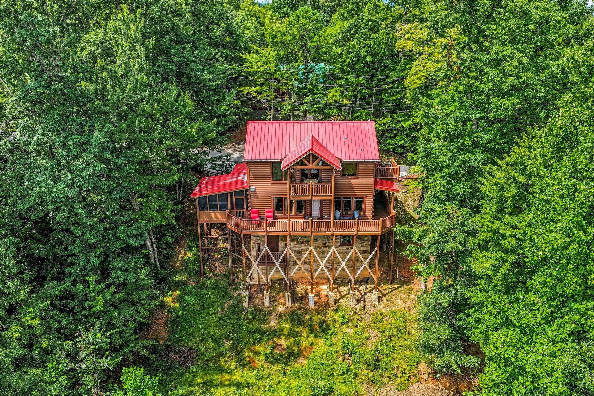 This charming log cabin sits nestled in the lush forest canopy on elevated stilts, featuring a striking red roof and wraparound deck for mountain living.