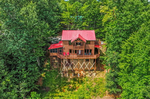 This charming log cabin sits nestled in the lush forest canopy on elevated stilts, featuring a striking red roof and wraparound deck for mountain living.