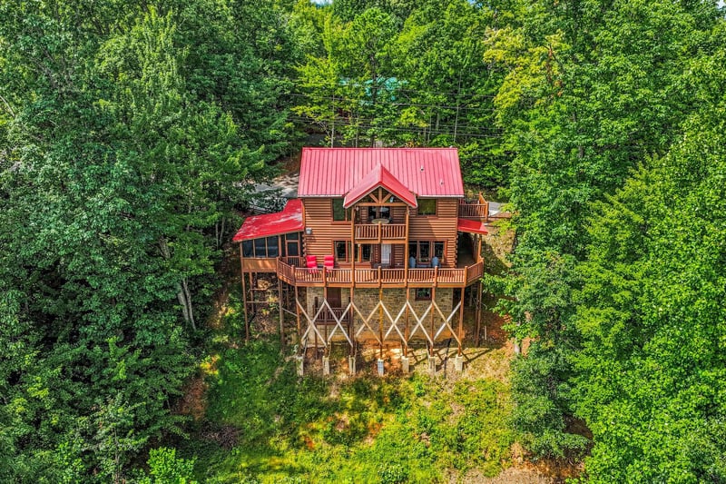This charming log cabin sits nestled in the lush forest canopy on elevated stilts, featuring a striking red roof and wraparound deck for mountain living.