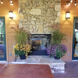 A striking stone fireplace anchors the covered outdoor patio, framed by vibrant fall planters and warm ambient lighting—a welcoming entrance to your mountain retreat.