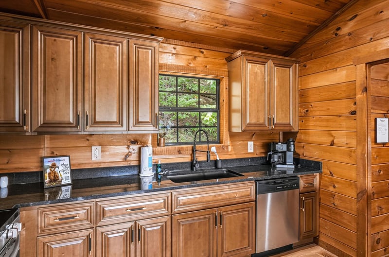 Your fully equipped kitchen features rich wood cabinetry, granite countertops, and modern stainless appliances, all bathed in natural light from the window overlooking the lush outdoors.