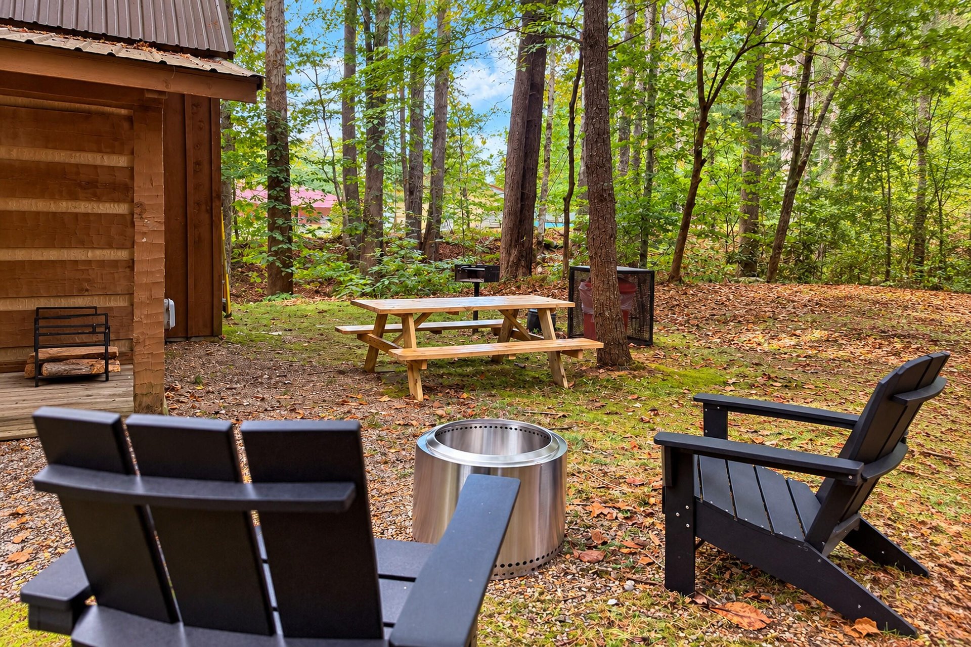 Your woodland retreat awaits with cozy outdoor seating around a sleek fire pit, plus a picnic table and BBQ area nestled among towering trees—perfect for fall evenings under the canopy.