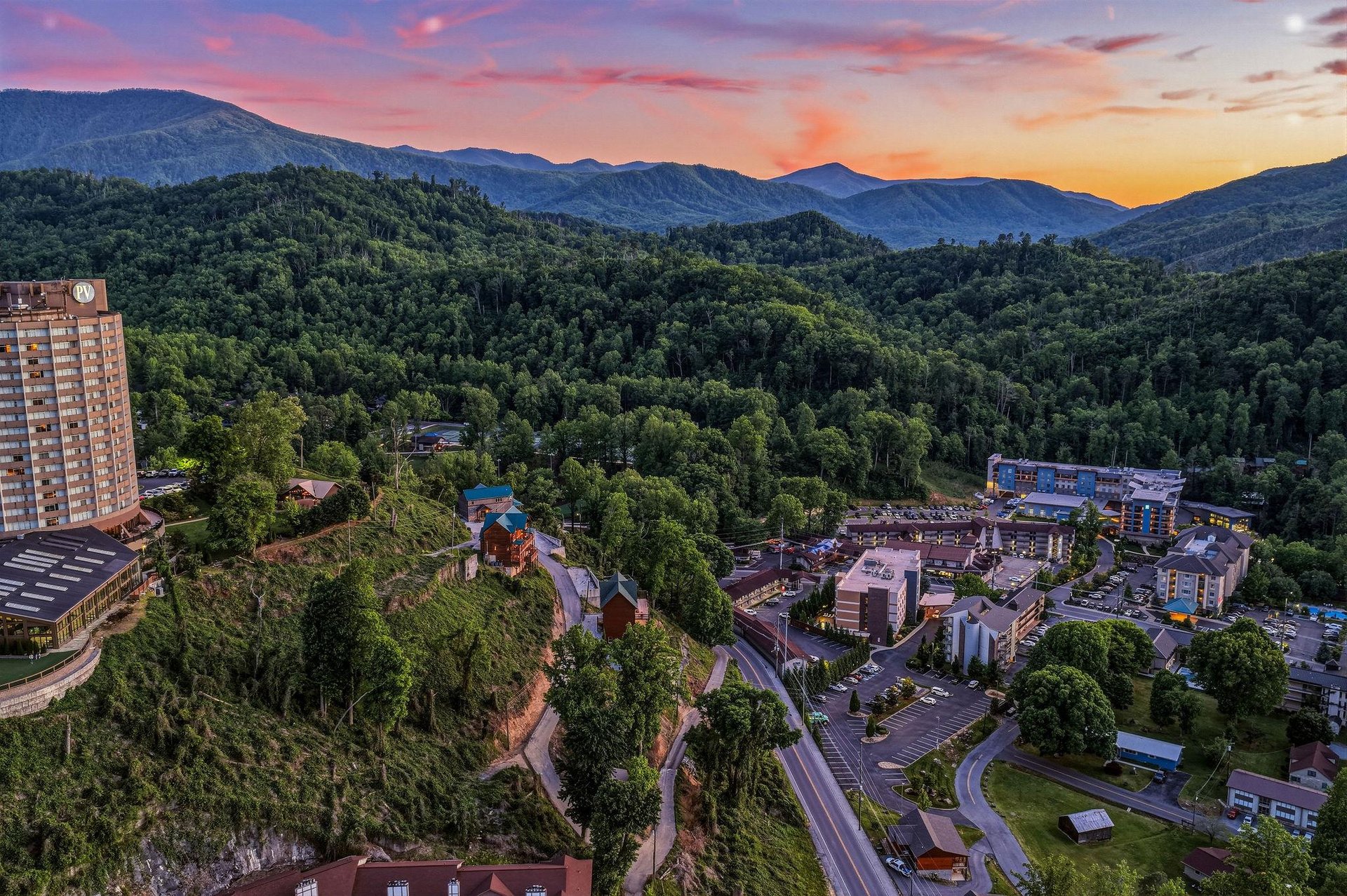 Aerial view of a mountain resort community nestled in forested hills, captured during sunset with dramatic pink and orange skies over the surrounding peaks.