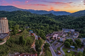 Aerial view of a mountain resort community nestled in forested hills, captured during sunset with dramatic pink and orange skies over the surrounding peaks.