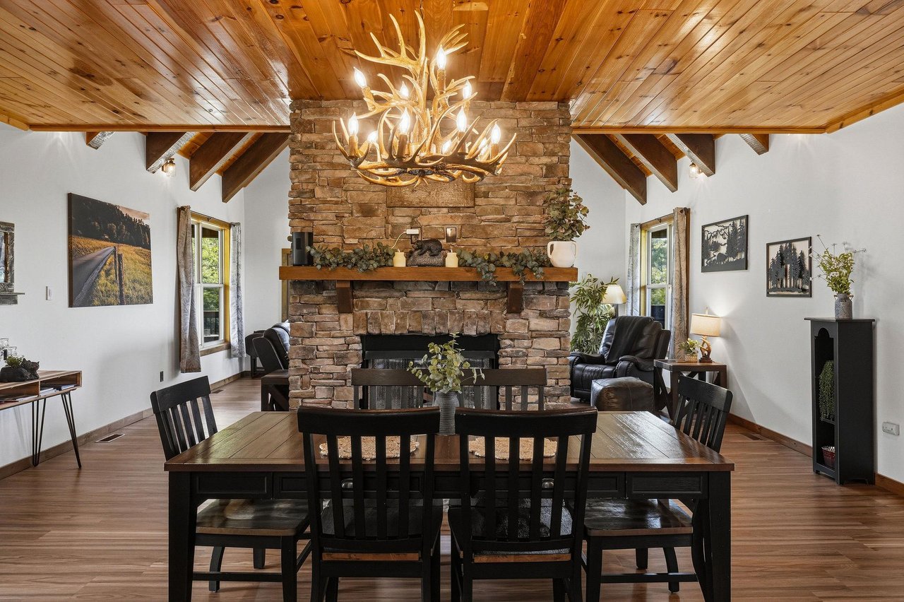 Gather around the rustic dining table under a stunning antler chandelier and warm woodbeamed ceilings, with the stone fireplace creating the perfect cozy centerpiece for memorable meals together.