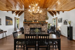 Gather around the rustic dining table under a stunning antler chandelier and warm woodbeamed ceilings, with the stone fireplace creating the perfect cozy centerpiece for memorable meals together.