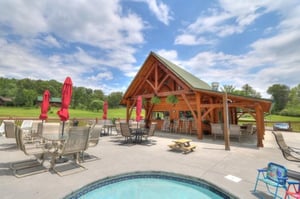 A covered outdoor pavilion features rustic log construction, offering shaded seating and dining areas beside the pool with mountain views beyond.
