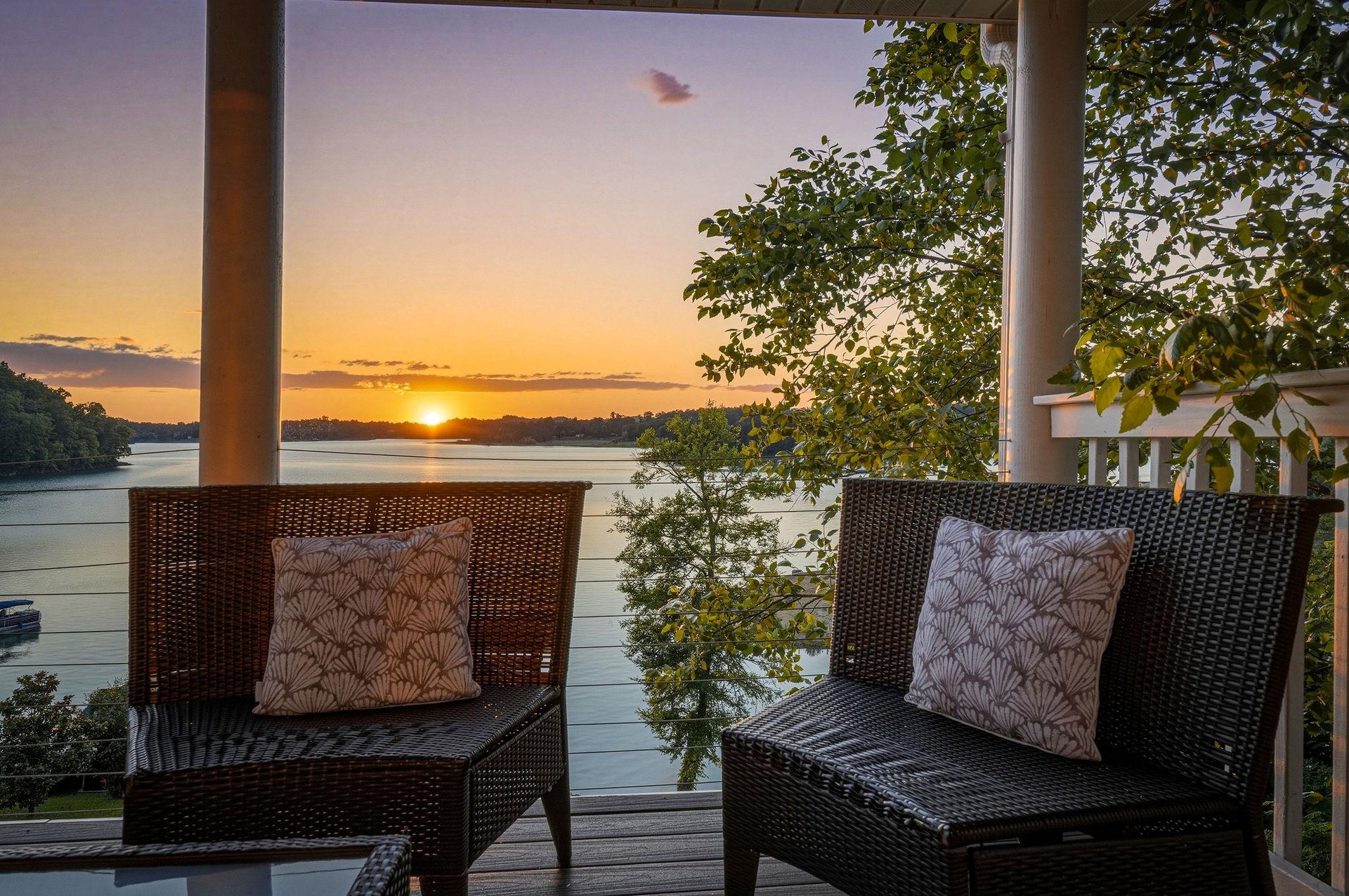 A covered terrace overlooks calm waters and lush greenery as the sun sets, framing two inviting wicker chairs with decorative cushions perfect for evening relaxation by the lake.