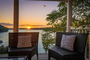 A covered terrace overlooks calm waters and lush greenery as the sun sets, framing two inviting wicker chairs with decorative cushions perfect for evening relaxation by the lake.