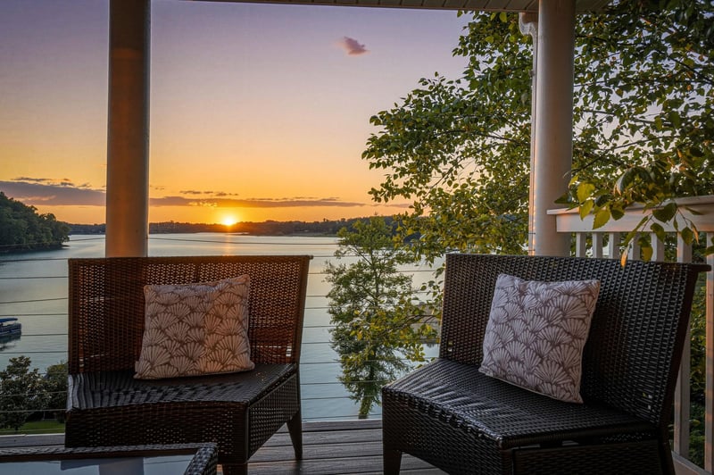 A covered terrace overlooks calm waters and lush greenery as the sun sets, framing two inviting wicker chairs with decorative cushions perfect for evening relaxation by the lake.