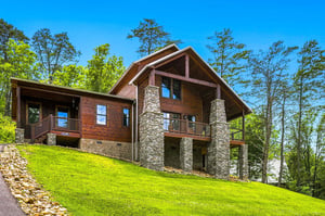 A striking mountain cabin with stone pillars and warm wood siding sits on a lush hillside surrounded by tall trees under a clear blue sky.