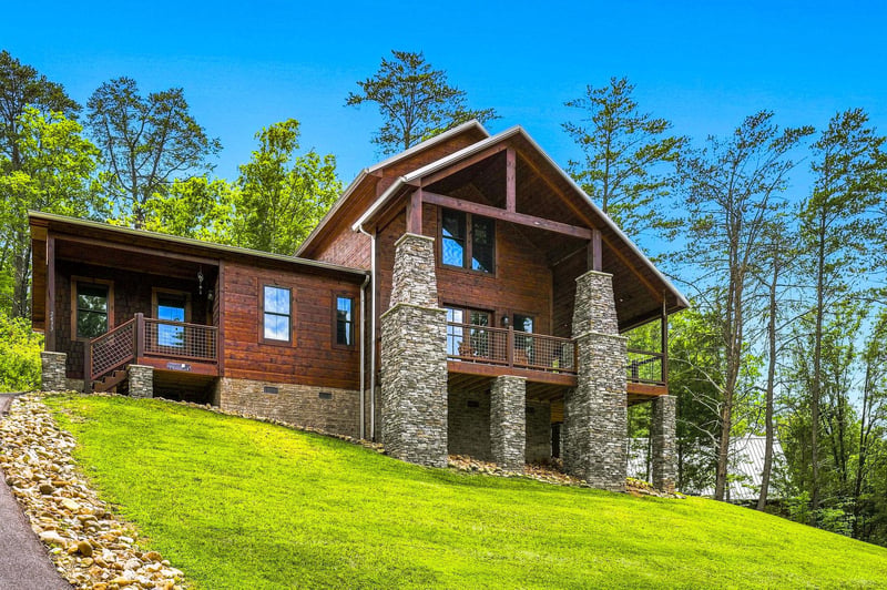 A striking mountain cabin with stone pillars and warm wood siding sits on a lush hillside surrounded by tall trees under a clear blue sky.