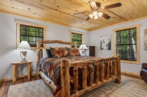 Your cozy mountain bedroom features a handcrafted log bed under a pine ceiling, with a ceiling fan to keep you comfortable and forest views through every window.