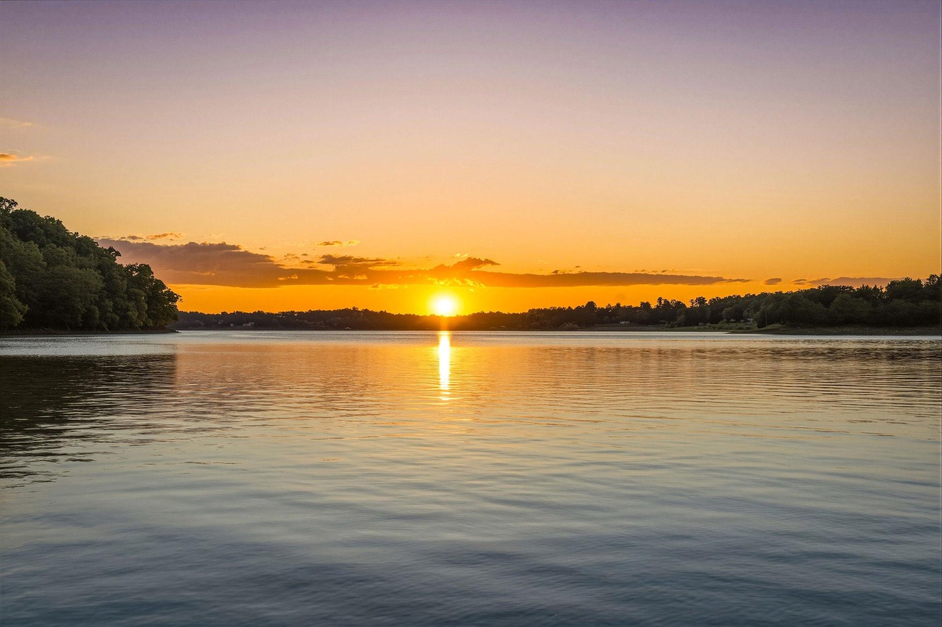 A stunning sunset paints the sky in vibrant golds and oranges, casting a warm glow over the tranquil lake surrounded by lush trees and natural beauty.