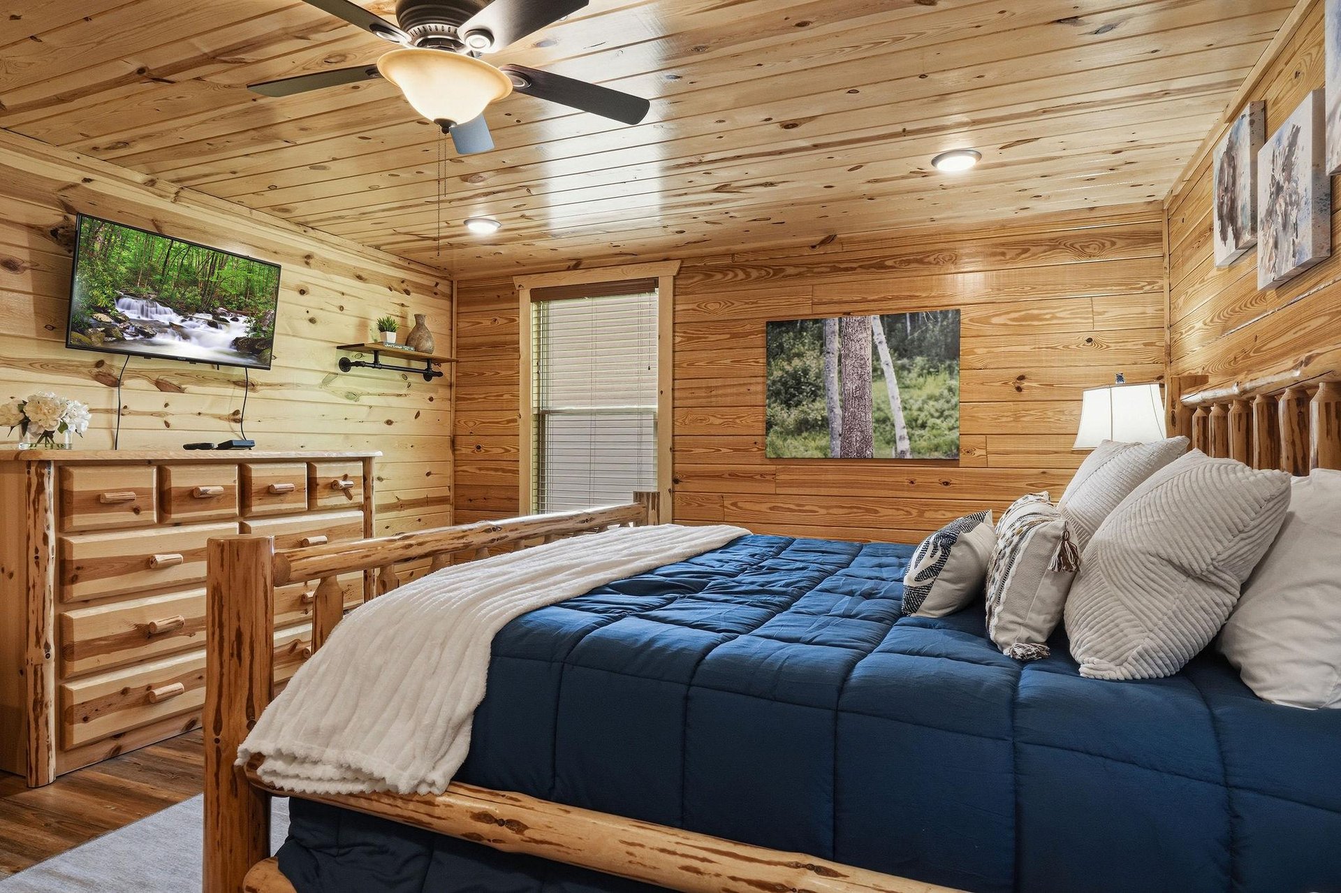 Your cozy retreat features handcrafted log furnishings, a plush bed dressed in soft linens, a mounted TV for relaxation, and warm woodpaneled walls adorned with natureinspired artwork.