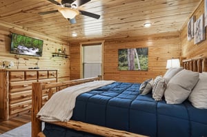 Your cozy retreat features handcrafted log furnishings, a plush bed dressed in soft linens, a mounted TV for relaxation, and warm woodpaneled walls adorned with natureinspired artwork.