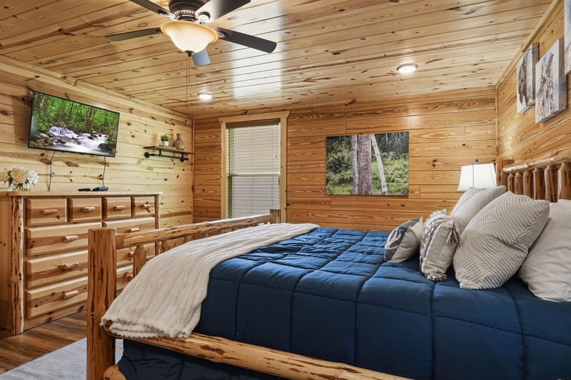 Your cozy retreat features handcrafted log furnishings, a plush bed dressed in soft linens, a mounted TV for relaxation, and warm woodpaneled walls adorned with natureinspired artwork.