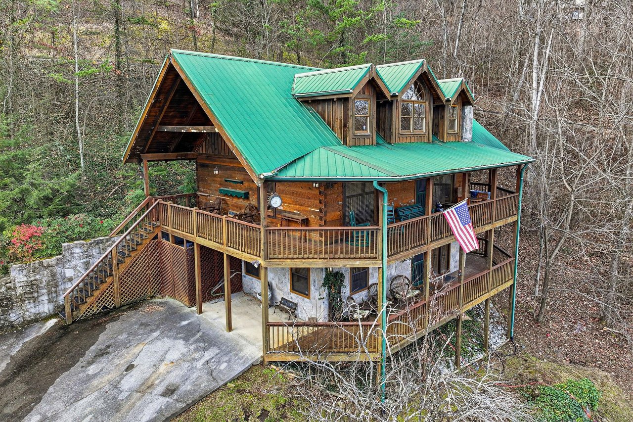 Rustic mountain cabin with green metal roof nestled in wooded hillside, featuring wraparound decks and forest surroundings.