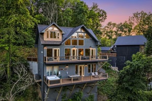 A stunning modern mountain retreat nestled among lush trees, featuring expansive windows and spacious decks to frame breathtaking sunset views over the forested hillside.