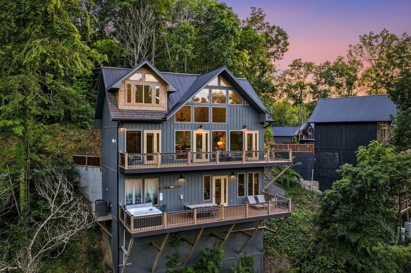 A stunning modern mountain retreat nestled among lush trees, featuring expansive windows and spacious decks to frame breathtaking sunset views over the forested hillside.