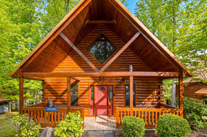A stunning log cabin nestled among towering trees, featuring a welcoming red door and covered porch perfect for morning coffee.