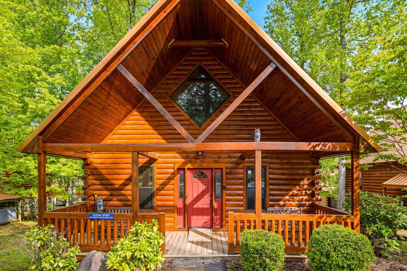 A stunning log cabin nestled among towering trees, featuring a welcoming red door and covered porch perfect for morning coffee.