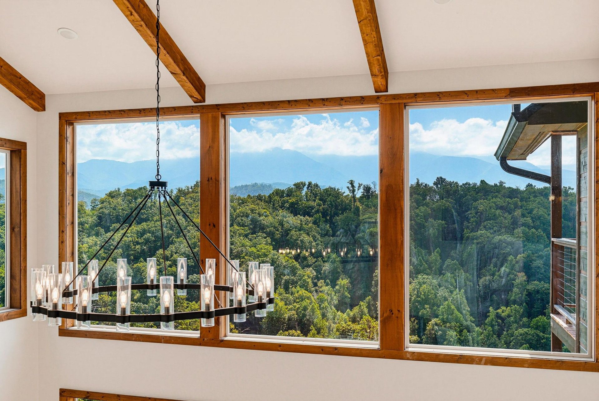 Sunlight streams through large windows framing mountain peaks and lush forests, while exposed wooden beams add warmth overhead. Imagine mornings here with coffee in hand, soaking in this serene natura