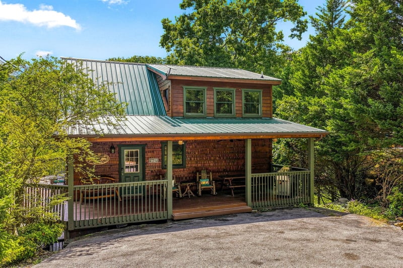 A charming twostory cabin nestled among towering trees, featuring rustic wood siding and a welcoming covered front porch with seating, offering a peaceful wooded retreat.
