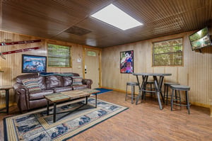 Your cozy basement retreat features warm wood paneling, comfortable leather seating, and natural light from a skylight above. Perfect for relaxing evenings with friends around the hightop table.