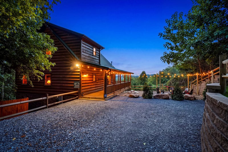 This charming log cabin property sits nestled among trees with welcoming outdoor lighting and a spacious gravel parking area, offering a cozy mountain retreat surrounded by natural beauty.