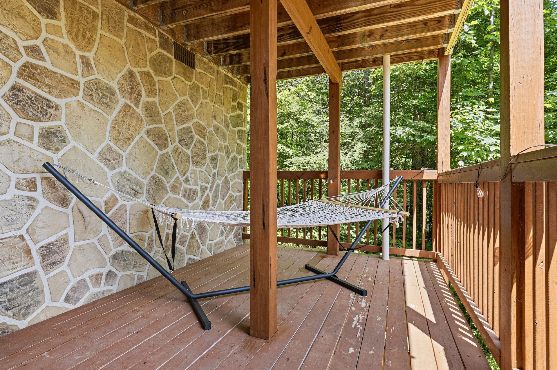 Swing gently in your hammock beneath a rustic wooden ceiling, stone wall at your back, and lush green forest just beyond the railing.