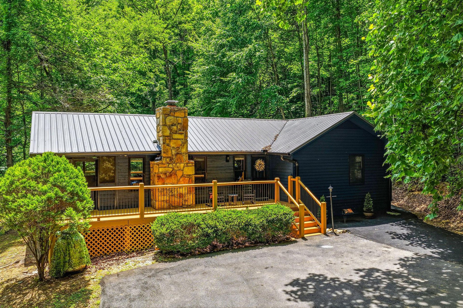 Charming woodland cabin with stone chimney and spacious wraparound deck, nestled among mature trees in a peaceful forested setting.